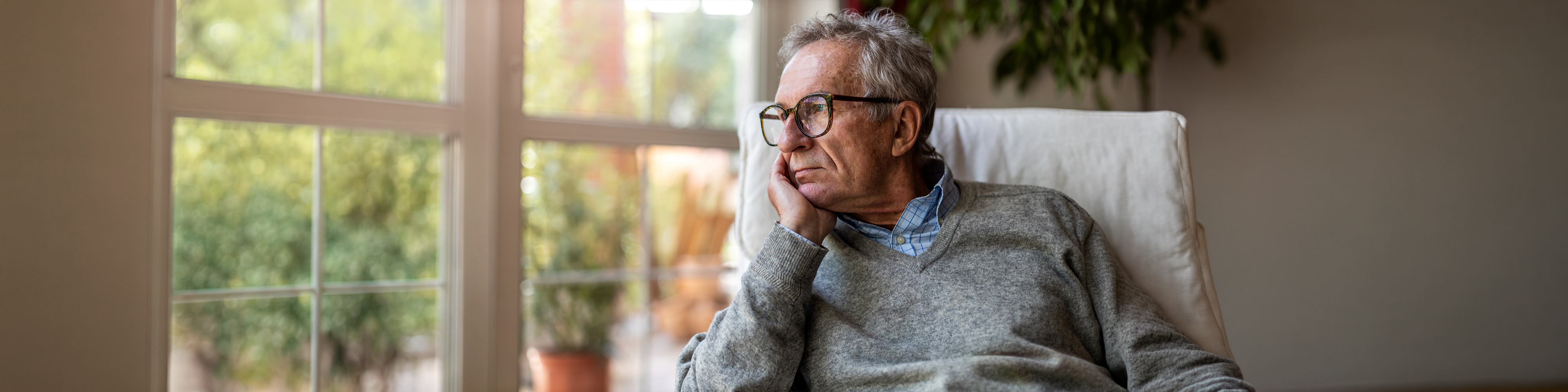 Elderly man with sad face thinking by the window in his living room 